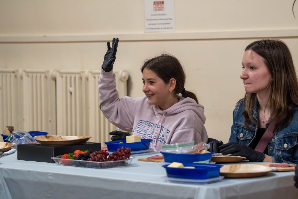 Two girls at a table with charcuterie supplies, one raising her hand