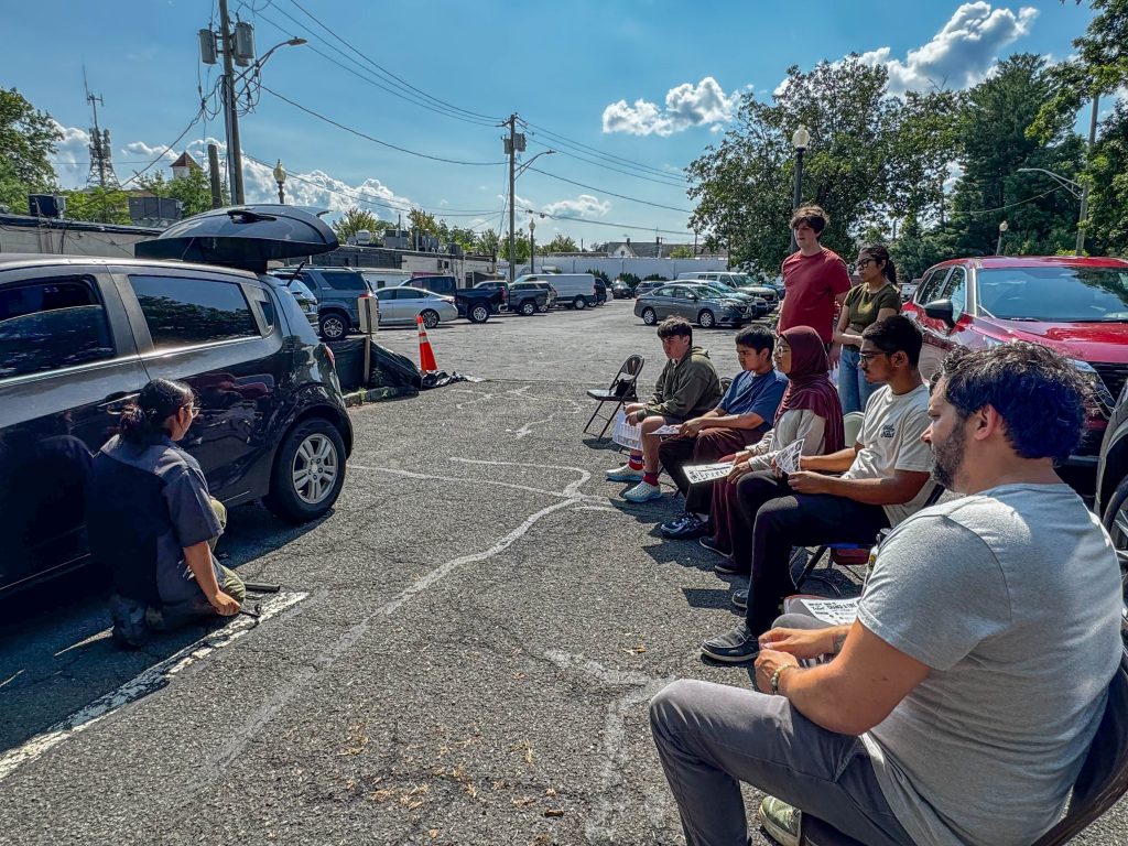 A group of people observing a car maintenance demonstration outdoors