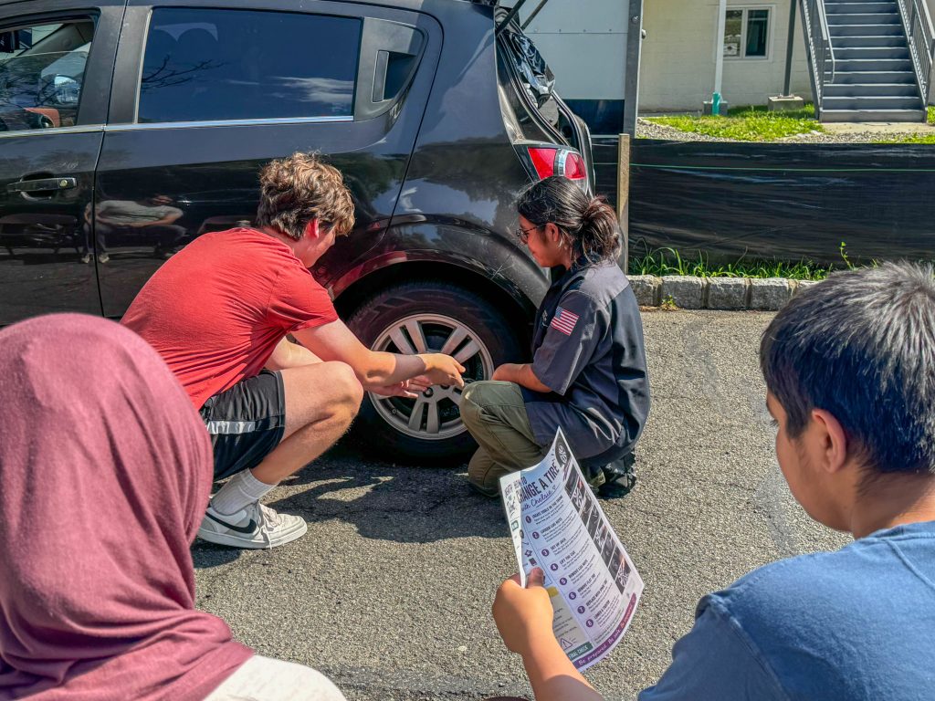 Two individuals work on a car tire while others observe nearby