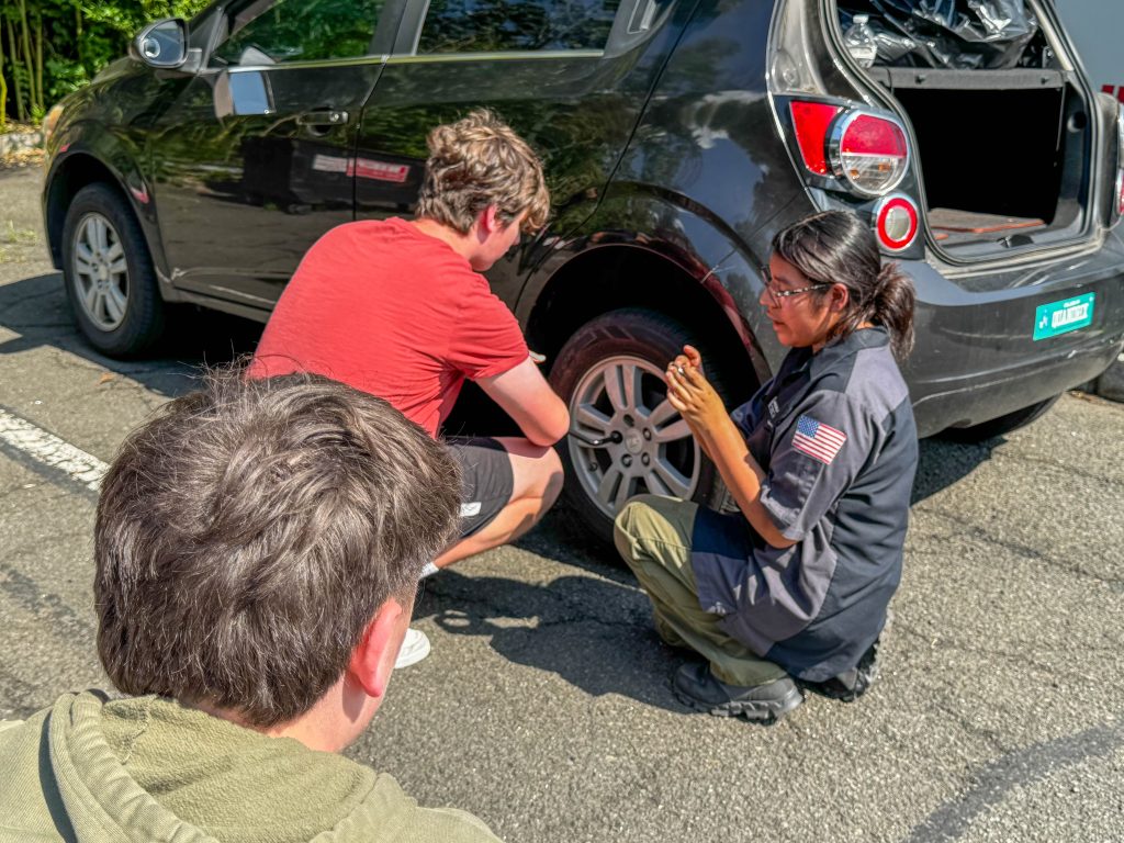 A woman teaches two young men how to change a tire